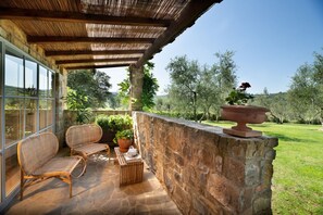 A covered patio with wicker furniture, a stone wall, and a view of the surrounding landscape. - Charming Villa with Private Pool immersed in the greenery of Chianti (Panzano In Chianti)