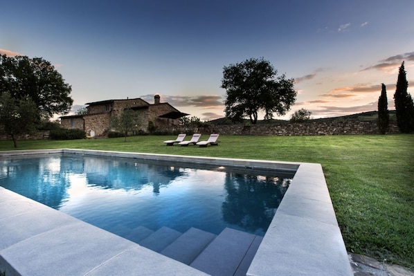 A swimming pool with lounge chairs, a stone building, and a grassy area with trees.