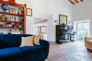 A living room with a blue sofa, a bookshelf, a piano, and a view through a window. - Charming Villa with Private Pool immersed in the greenery of Chianti (Panzano In Chianti)