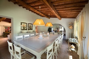 A spacious dining area with a long table, white chairs, and hanging lamps. There is a kitchen visible in the background.