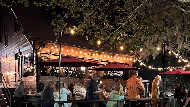 An outdoor seating area with tables and chairs, string lights overhead, and a signboard listing menu items.