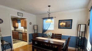 A dining area with a wooden table, chairs, and a bench. There is a window with blue curtains and a hanging light fixture. A kitchen is visible in the background.