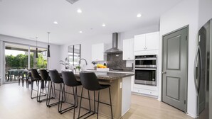 A modern kitchen with a central island, bar stools, a built-in oven, and a large door. - Paseo Palms, Modern 6br home 5min to Polo Grounds (Coachella)
