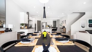 A modern dining area with a long table set for a meal, surrounded by chairs, and a kitchen in the background. - Paseo Palms, Modern 6br home 5min to Polo Grounds (Coachella)