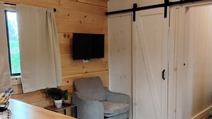 A cozy room with wooden walls, a grey armchair, a dining table with bar stools, a TV mounted on the wall, and a sliding door closet. - Serenity at Nottely River (Murphy)