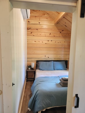 Quarto com teto de madeira, cama com roupa de cama azul, mesa de cabeceira com abajur e criado-mudo com uma planta.