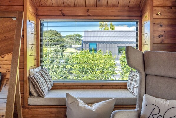 A wooden cabin interior with a window seat, a cushioned bench, and a view of a modern building outside.