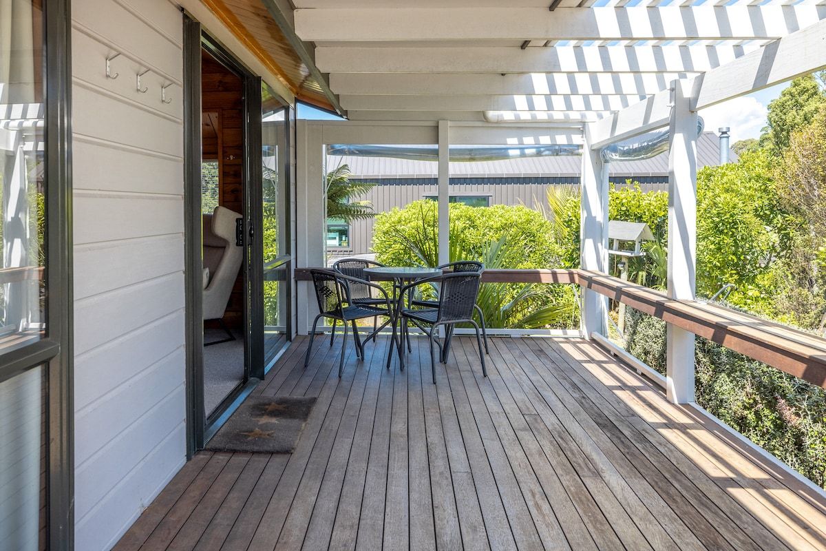A wooden deck with a table and chairs, a pergola, and a view of greenery.