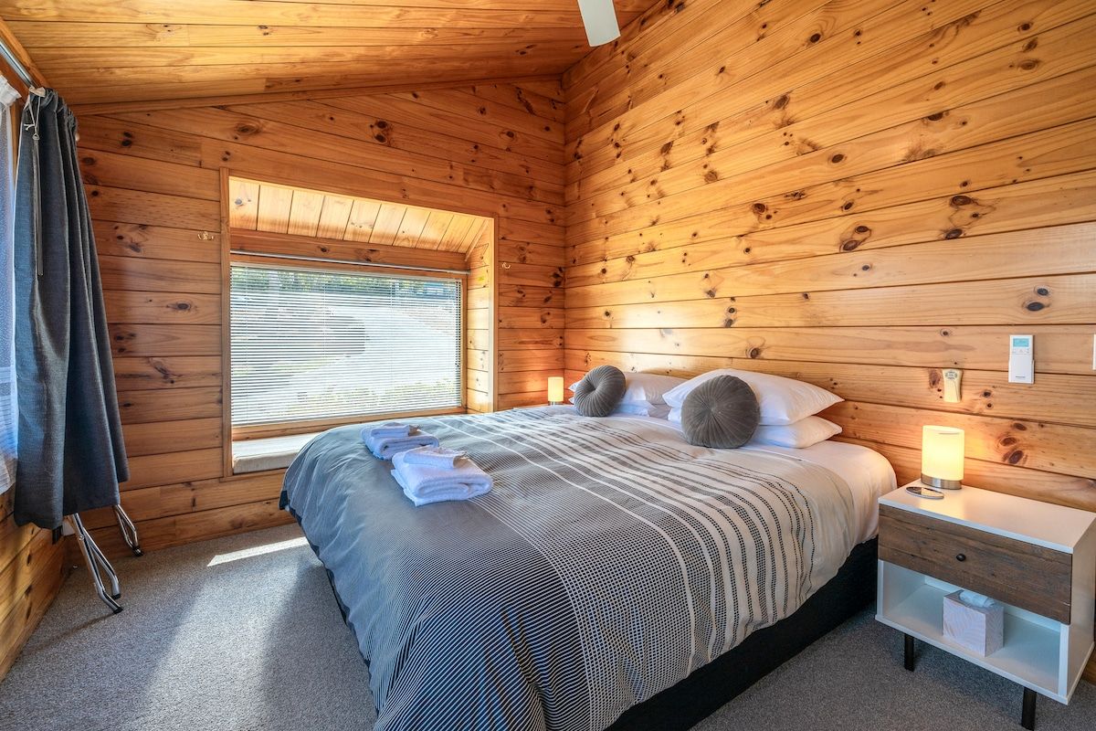 Una habitación con paneles de madera, una cama, un ventanal, una mesita de noche y una lámpara.