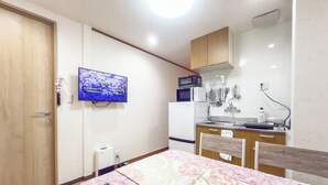 A compact kitchen with a microwave, refrigerator, and a wall-mounted TV. A remote control is on a table with a floral tablecloth.