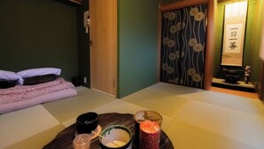 A traditional Japanese room with tatami flooring, a bed with a pink blanket, a small table with a bowl and a glass of drink, and a sliding door with floral patterns.