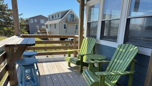 Una terraza de madera con sillas Adirondack verdes y una pequeña mesa, con vista a una zona residencial con casas y un cielo despejado.