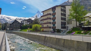 A hotel with multiple floors, a river in the foreground, and mountains in the background.