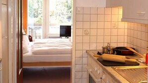 A bedroom with a bed, a TV, and a window with a view of greenery. The kitchen has white cabinets, a stove, and a sink.