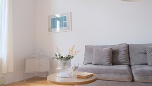 Een moderne woonkamer met een grijze bank, een ronde houten tafel met een bloevas, een wit kastje en een lijst met een afbeelding aan de muur.