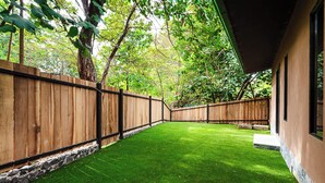 A fenced backyard with artificial grass, a stone wall, and a building with windows. - Private Tropical Villa with Backyard • Pool • Fast WiFi • Self Check-In (Provincia de Guanacaste)