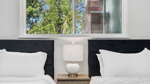 A hotel room with two beds, a nightstand with a lamp, and a window with a view of greenery.