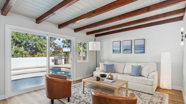 A modern living room with a white sofa, brown leather chairs, a wooden coffee table, and a large window overlooking a pool and greenery.