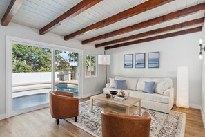 A modern living room with a white sofa, brown leather chairs, a wooden coffee table, and a large window overlooking a pool and greenery.