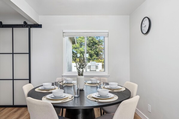 A dining area with a round table set for four, a window with a view of trees, and a wall-mounted clock.