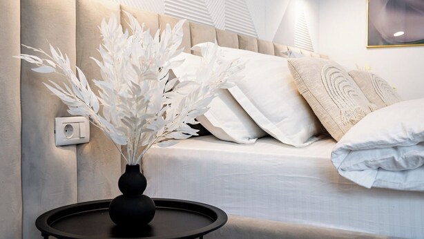 A modern bedroom with a beige sofa, white pillows, and a vase with white flowers on a small table.