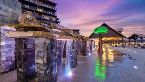 Una estructura de cascada de piedra con un paraguas iluminado en verde en primer plano, un edificio con balcones y una zona de estar cubierta al fondo.