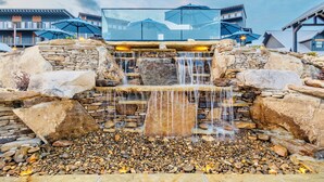Una cascada con un recipiente de piedra y guijarros, rodeada de rocas grandes y un edificio moderno con balcones y sombrillas.