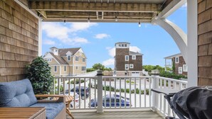 A wooden deck with a wicker sofa and grill, overlooking a residential area with houses and parked cars.