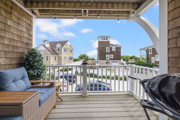A wooden deck with a wicker sofa and grill, overlooking a residential area with houses and parked cars.