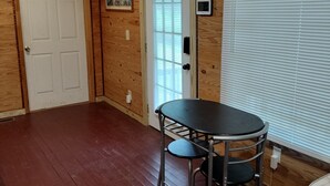 A room with wooden walls and a wooden ceiling, a white door, a window with blinds, a round table with two chairs, and a small wall-mounted TV. - 1-bedroom apartment in the rear suite of the Howard Finster Vision House Museum. (Summerville)