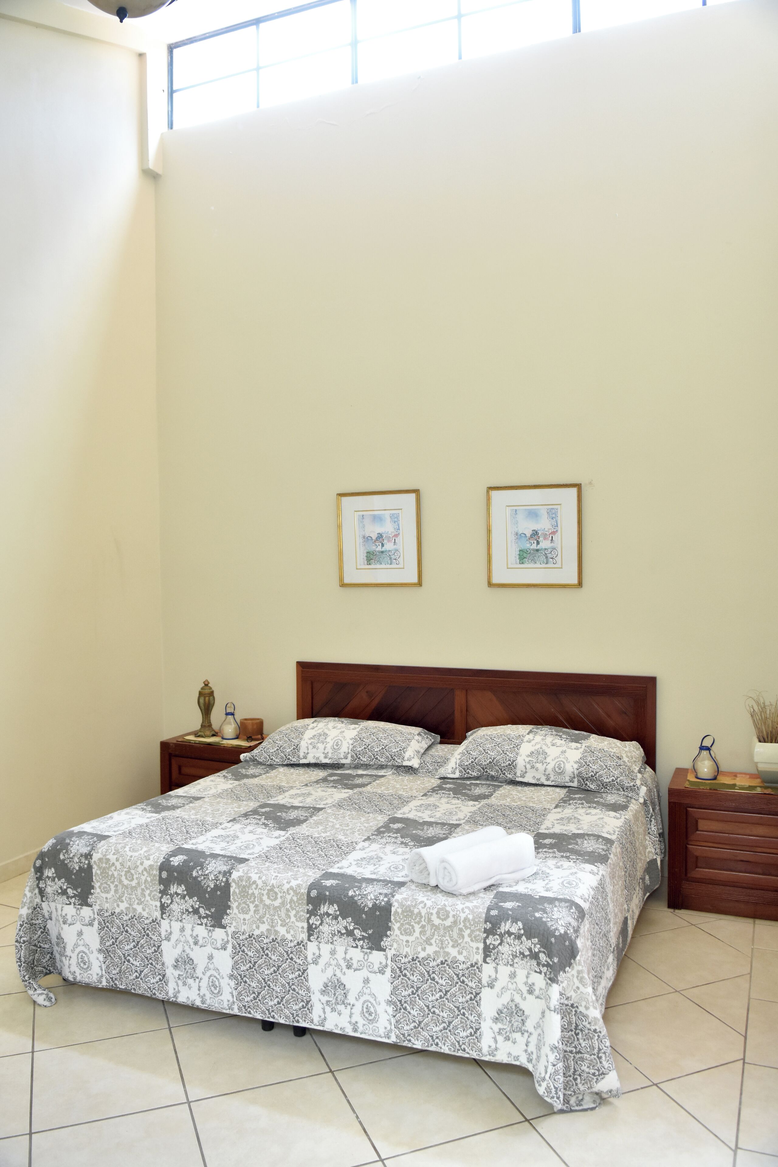 A bedroom with a large bed, two framed pictures on the wall, and a nightstand with a lamp.
