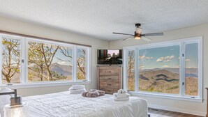 A bedroom with a large bed, two windows with mountain views, a ceiling fan, and a wooden dresser.