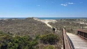 Una pasarela de madera que lleva a una playa con vegetación escasa y un cielo azul despejado.