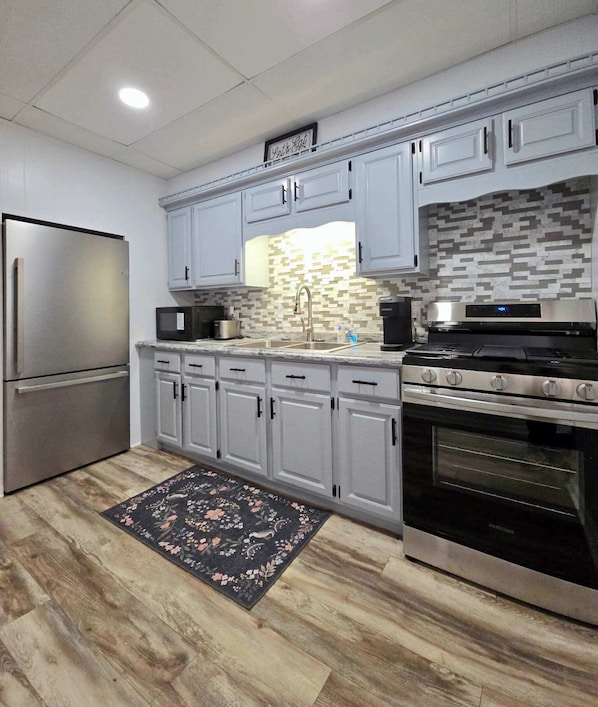A modern kitchen with grey cabinets, a stainless steel refrigerator, a microwave, a sink, a stove, and a patterned backsplash.