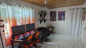 Ein Zimmer mit Holzboden, einem Esstisch mit zwei Computermonitoren, orangefarbenen Stühlen, einem schwarzen Sofa und einem Fliesenboden.