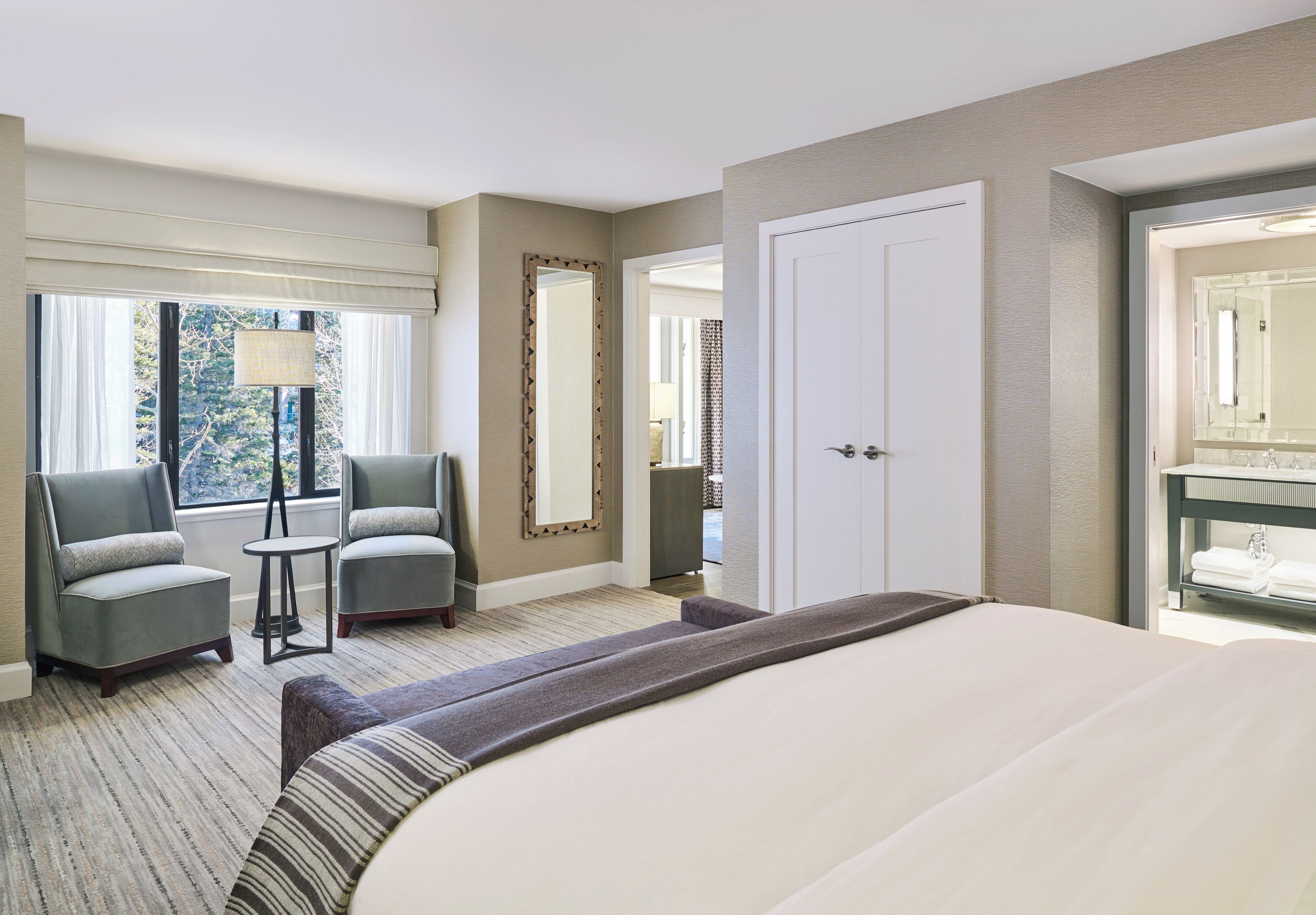 Une chambre d’hôtel comprenant un lit, deux fauteuils, une petite table, un miroir et une porte donnant sur une salle de bain.