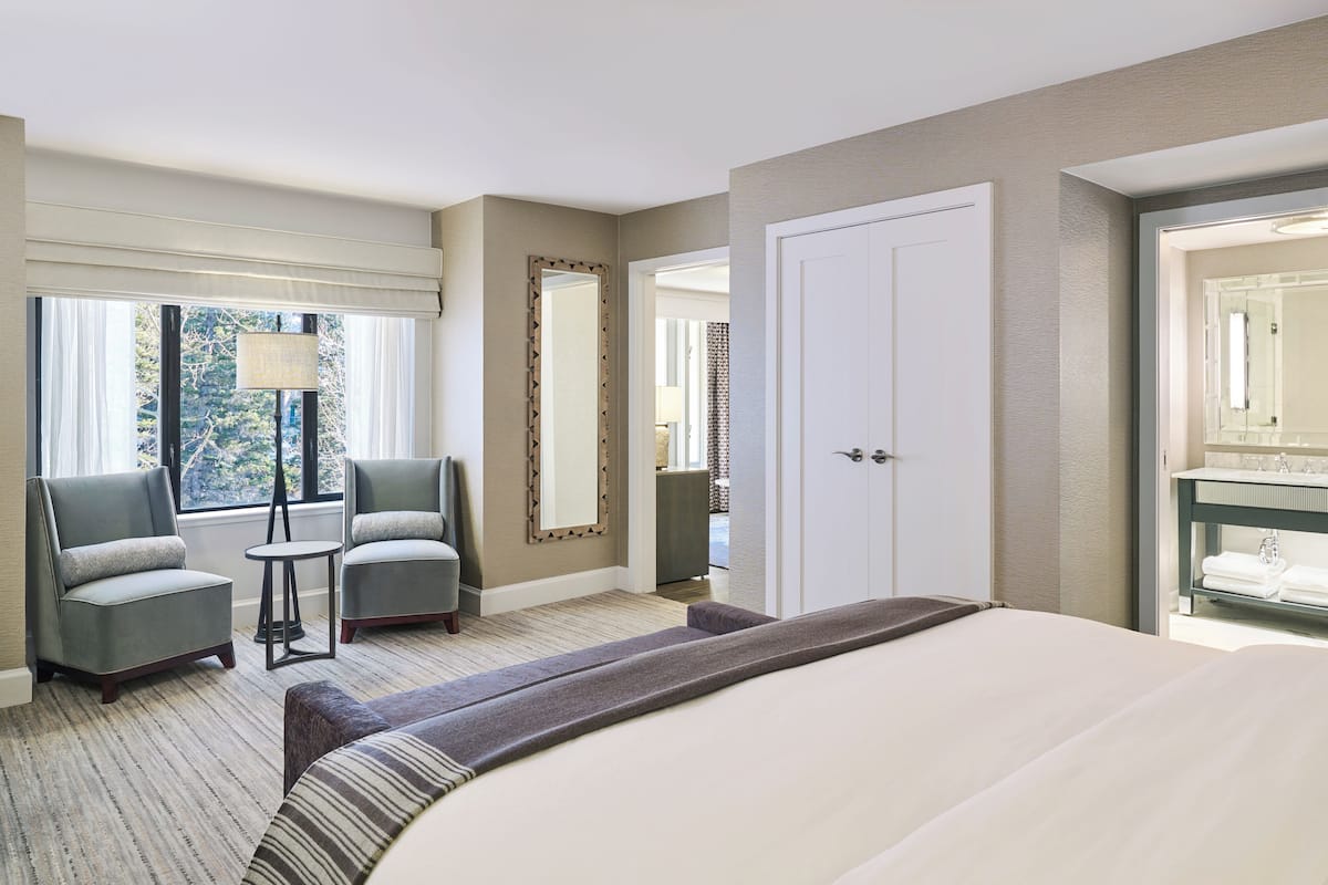 Une chambre d’hôtel comprenant un lit, deux fauteuils, une petite table, un miroir et une porte donnant sur une salle de bain.
