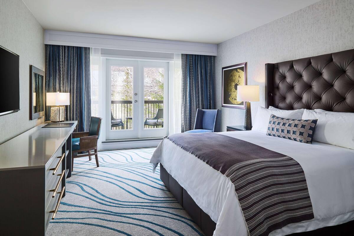 Une chambre d’hôtel avec un lit, un bureau, une chaise, une télévision et un balcon avec des rideaux.