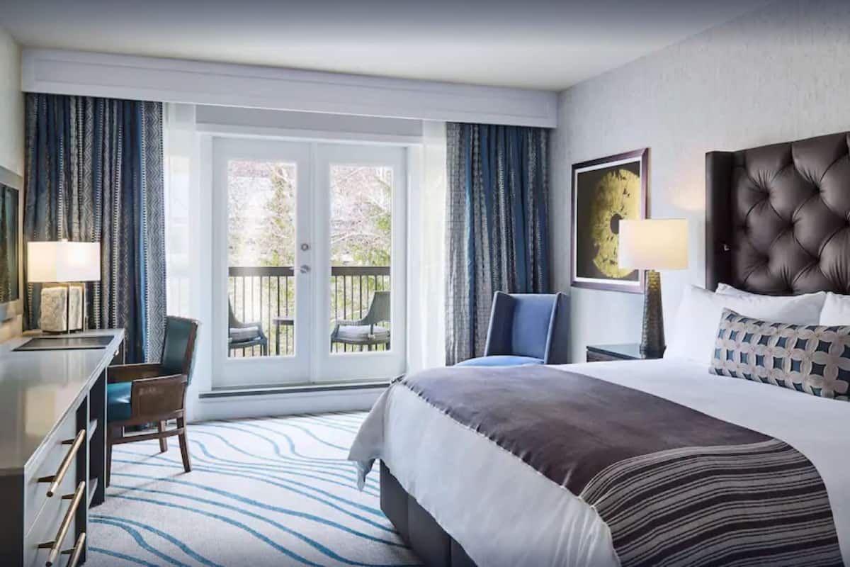 A hotel room with a bed, a desk, a chair, a balcony with a view, and a window with curtains.