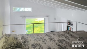 A bedroom with a large window, a bed with a patterned bedspread, a small balcony, and a view of a green landscape. - Spinola Beachfront Bungalows - La Palmera (Sanlúcar de Barrameda)
