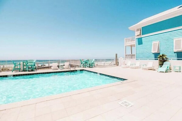 A pool area with a clear view of the ocean, a blue building with a balcony, and several lounge chairs.