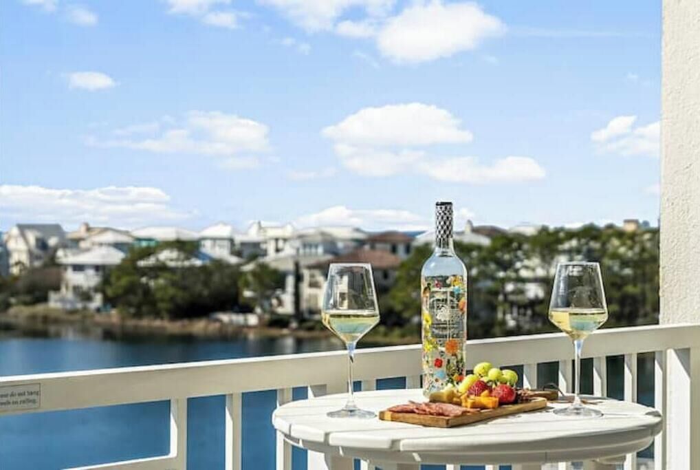 A table with two glasses of white wine, a bottle of wine, and a fruit platter on a balcony overlooking a body of water and buildings.