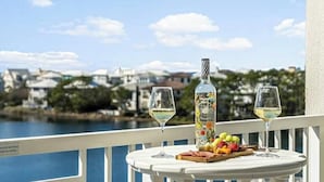 Et bord med to glas hvidvin, en vinflaske og et frugtfat på en balkon med udsigt over vand og bygninger.