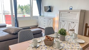 Una sala moderna con un sofá, una mesa de comedor con platos, tazas y una cesta de pan. Un televisor está montado en la pared, y hay una ventana con cortinas.