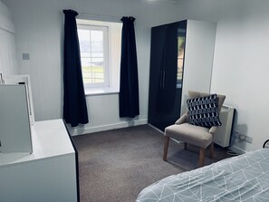 A room with a bed, a chair with a patterned pillow, a window with curtains, and a radiator.