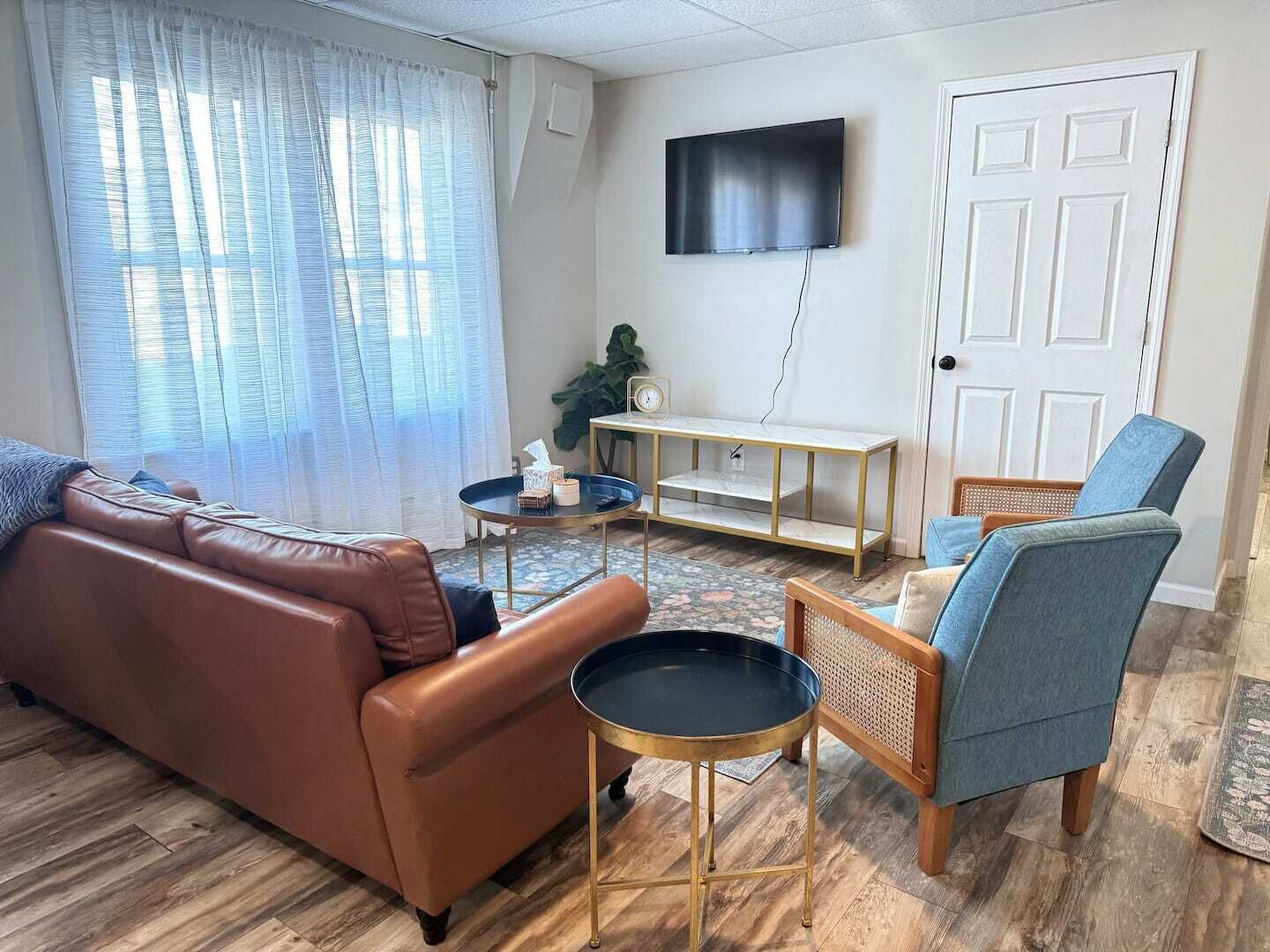 Una sala de estar con un sofá de cuero marrón, un sillón azul, una pequeña mesa redonda, un televisor montado en la pared y una puerta de madera.