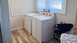 A laundry room with two washing machines, a small table with a chair, and a window with curtains.