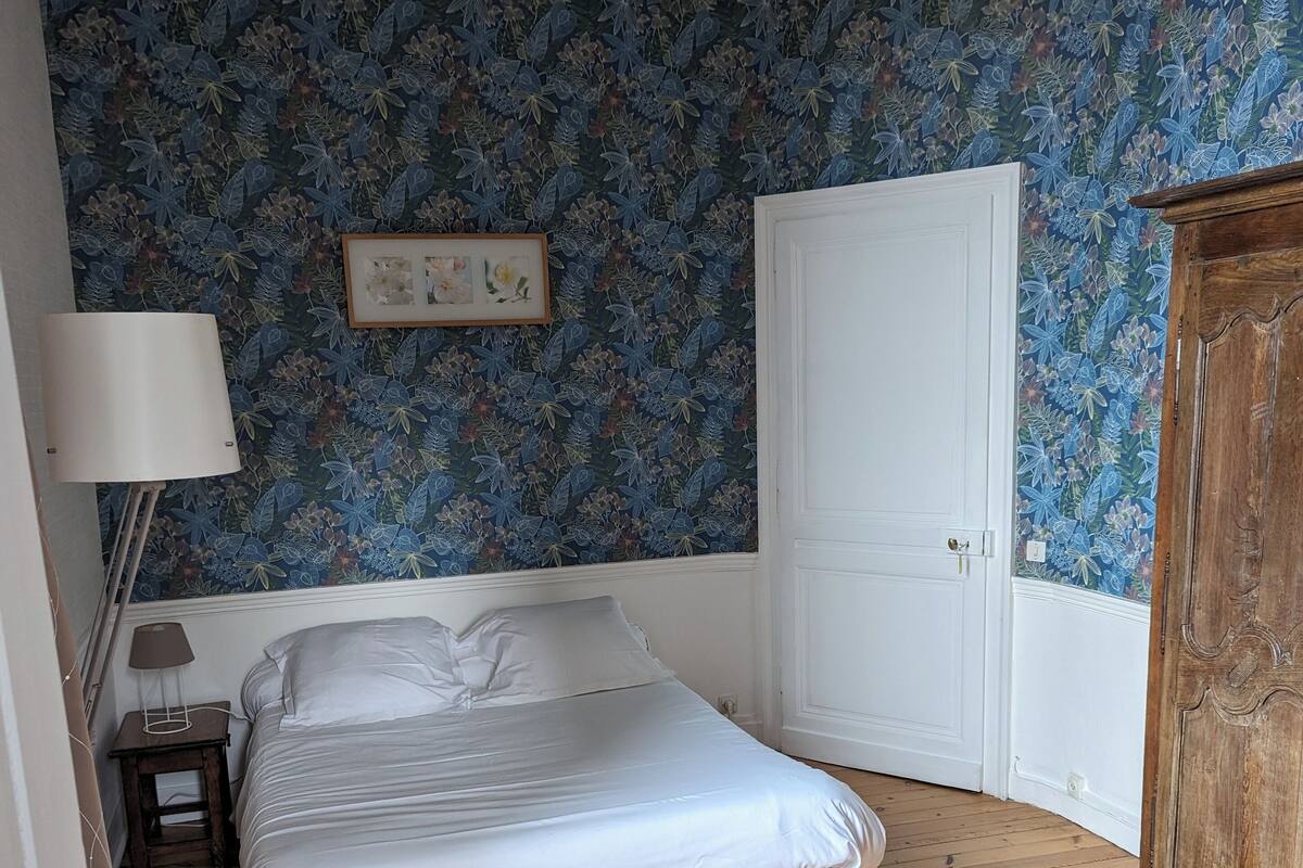 Une chambre à coucher avec un lit, un plancher en bois, un papier peint à motifs floraux, une porte blanche et une armoire en bois.