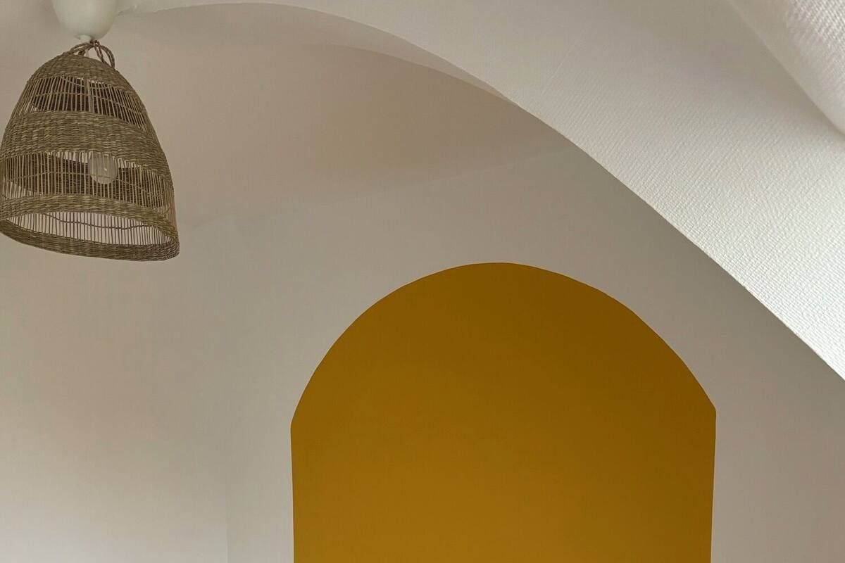 Une chambre à coucher avec un lit, deux tables de chevet, une lampe suspendue et un mur d’accent jaune.