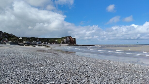 A pebble beach with a rocky cliff and houses in the background.